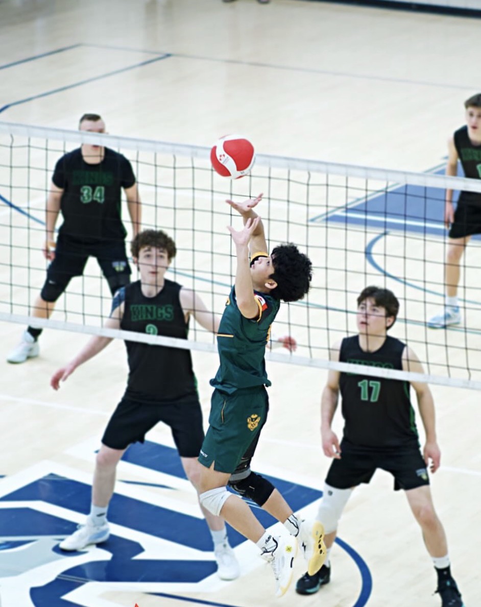 A setter in green jumping to set the ball at the net against three blockers in black